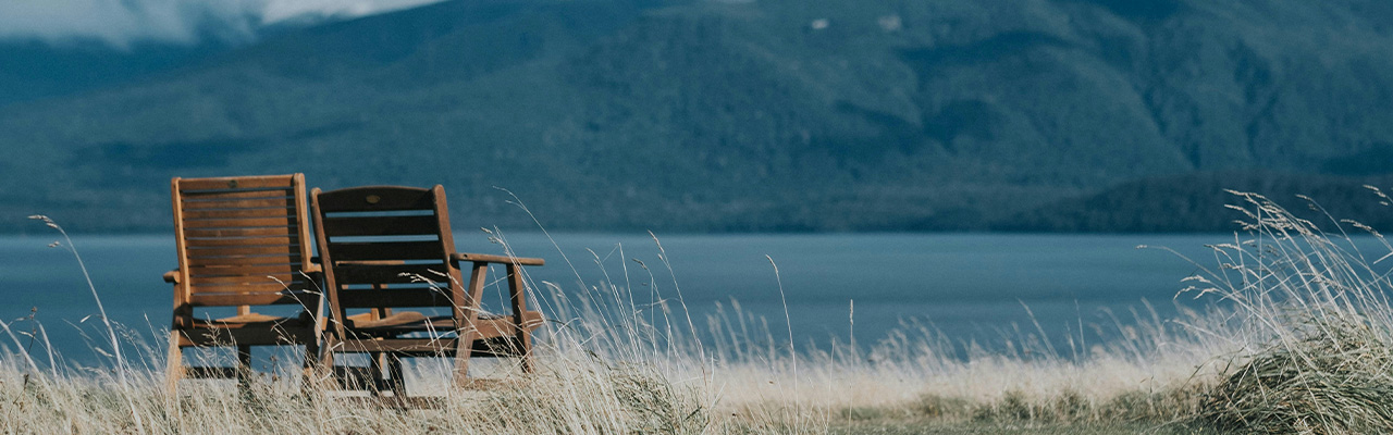 Two wooden Adirondack chairs in tall grass facing a lake and mountains
